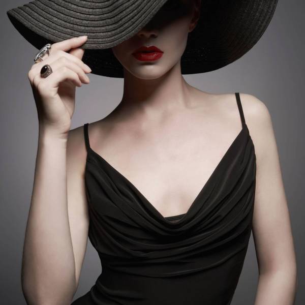 Fashion portrait of young sexy lady with beautiful black hat and evening dress. Stylish elegant woman with modern jewelry. Studio photo of pretty model on grey background.