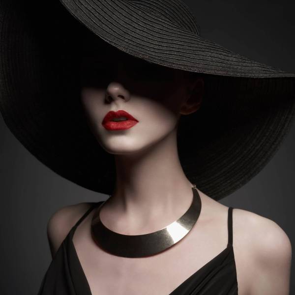 Fashion portrait of young sexy lady with beautiful black hat and evening dress. Stylish elegant woman with modern jewelry. Studio photo of pretty model on grey background.