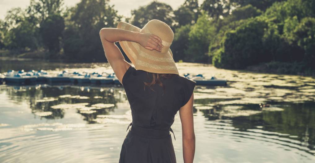 Vintage filtered shot of an elegant woman wearing a hat standing by a lake in a park at sunset