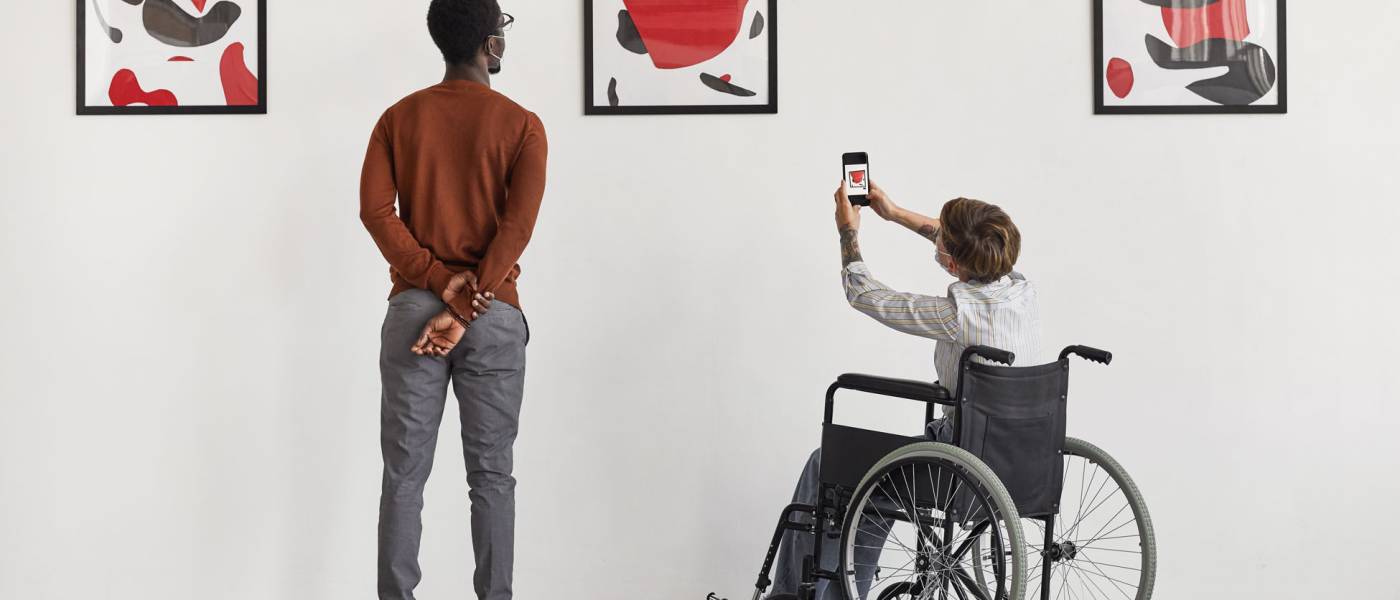 Back view portrait of young woman using wheelchair taking photo of artwork while visiting accessible museum, copy space