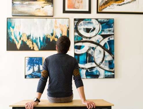 Hispanic male visitor looking reflective while sitting on a bench and admiring the various paintings on the wall of an art gallery