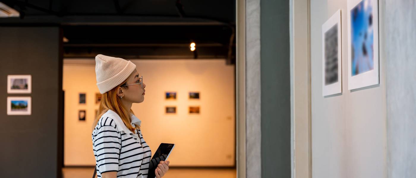 Young person at photo frame hold digital book leaning against at show exhibit artwork gallery, Asian woman holding tablet at art gallery collection in front framed paintings looking pictures on wall