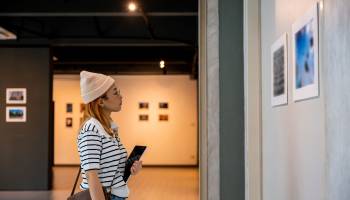 Young person at photo frame hold digital book leaning against at show exhibit artwork gallery, Asian woman holding tablet at art gallery collection in front framed paintings looking pictures on wall