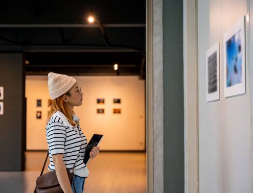 Young person at photo frame hold digital book leaning against at show exhibit artwork gallery, Asian woman holding tablet at art gallery collection in front framed paintings looking pictures on wall