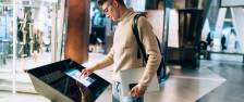 Side view of handsome young man in glasses and with backpack standing with gadgets in hands while using information panel in shopping centre